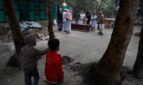 Two small children look on as people gather round a coffin lying on the ground outside next to a house.