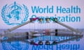 Photo of the World Health Organization (WHO) logo displayed on screen with medical syringes and vaccine vials