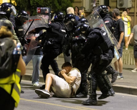 Police with helmets, batons and riot shields gather around a young man who is lying in the road; he has his hands up to his face