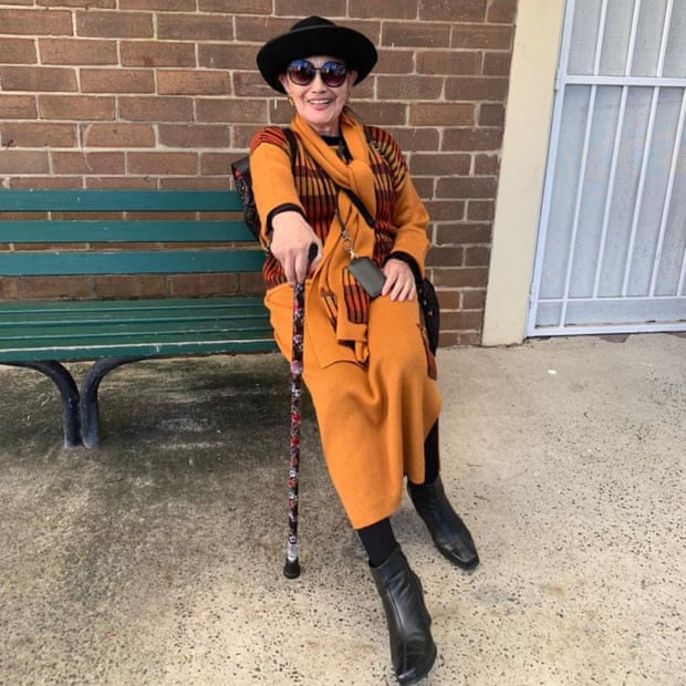 A woman sits on a green bench seat wearing orange clothing and holding a cane