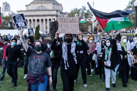 People dressed in black and white and wearing keffiyehs and holding signs and waving the Palestinian flag march across a green lawn.