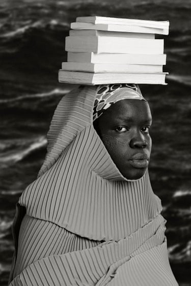 A woman wrapped in pleated fabric balances a stack of books on her head