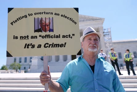 a man wearing a blue shirt and hat holds a sign that reads ‘plotting to overturn an election is not an official act, it’s a crime’