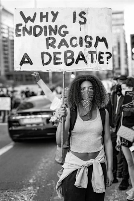 Darcy Bourne, with a bandanna tied over her nose and mouth, holds a handwritten sign saying Why is ending racism a debate?