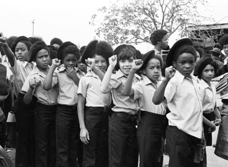 A line of Panther cubs in uniform stand facing the camera most with their fists raised in the Black Power salute.