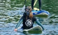 A young woman in a Surfers Against Sewage wetsuit paddling a surfboard while wearing a gas mask