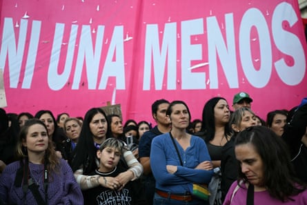 Latina women and a child stand listening to a speaker at a protest in front of a pink banner saying ‘Ni Una Menos’