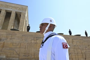 A soldier in a white uniform and helmet stands to attention in the foreground while others in black uniform stand on high wall behind him