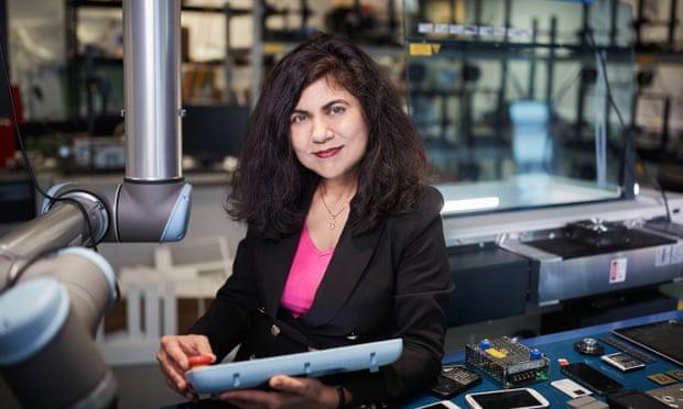Veena Sahajwalla is standing in a factory holding component. She is wearing a black suit and pink shirt and is looking at the camera