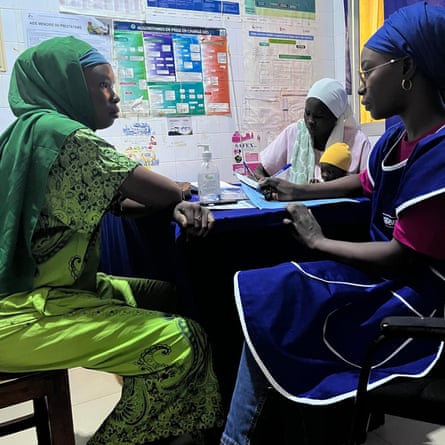 A young African woman in traditional headscarf and dress sits at a table in an office talking to a woman in jeans and gown, while another woman sits in the background