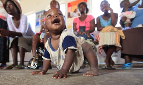 A smiling black baby crawls on the ground in front of a row of women sitting on chairs