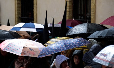 People shelter from the rain under umbrellas and some people are dressed in black pointed hoods
