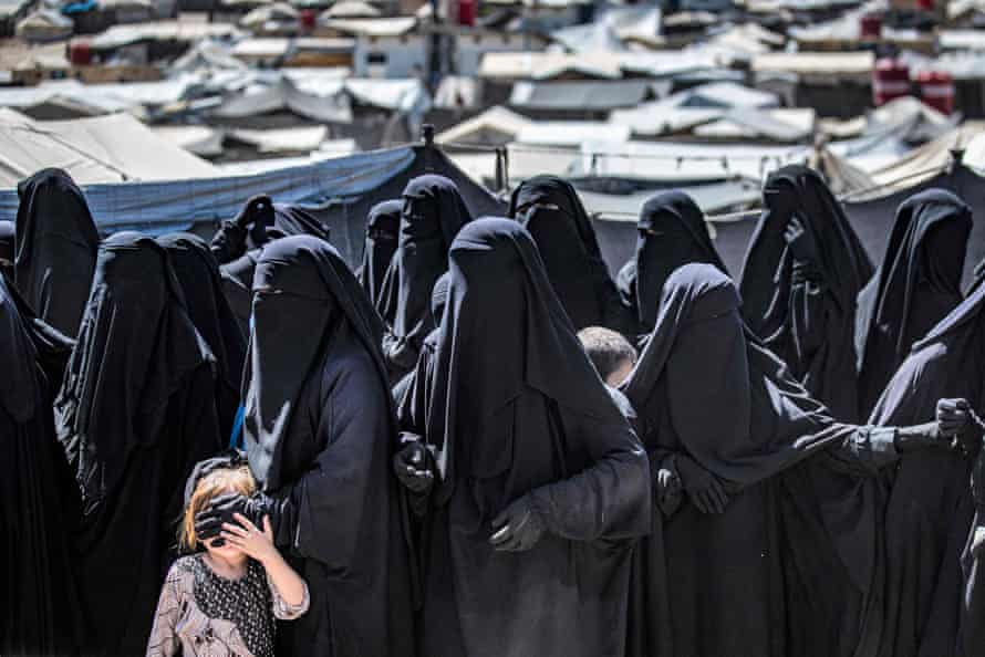 A group of women dressed in black robes, gloves and niqabs so their faces are completely covered. One woman covers the eyes of a child who is trying to look at the photographer