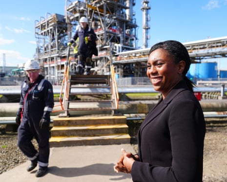 Kemi Badenoch in foreground with two workers in overalls and hard hats in background and industrial plant behind