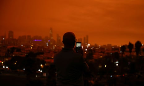 A man photographs Downtown San Francisco in Dolores Park under an orange sky darkened by smoke from California wildfires.