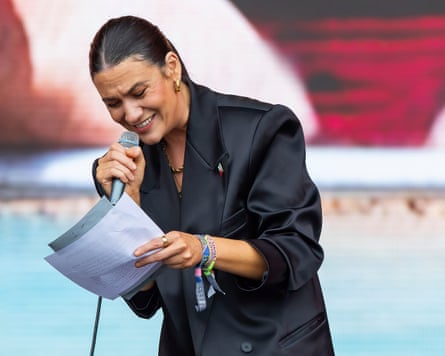 Nadine Shah reads from sheets of paper in her hand; she is wearing a black suit jacket and festival wristbands and has her dark hair tightly tied back.