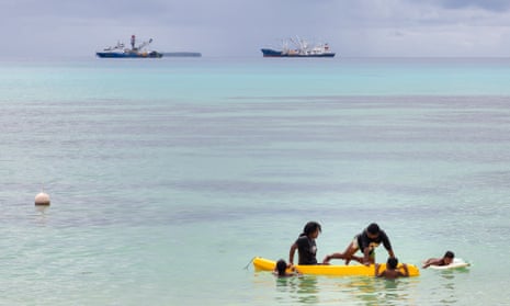 Children play in a small boat as large fishing boats sit offshore in Vanuatu