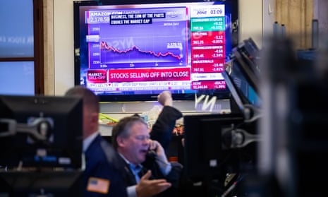 A large screen mounted on a wall shows Amazon's market loss. In the foreground, traders for the New York Stock Exchange sit at desks and talk on phones.
