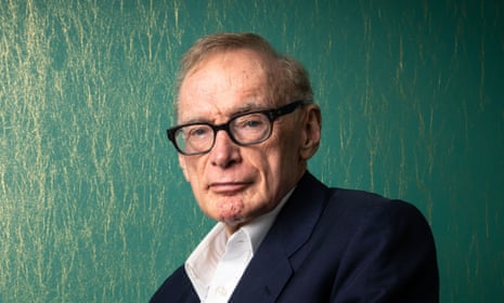A man with black glasses sits before a green background.