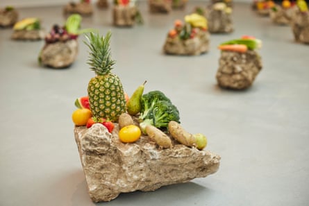 a large stone on which is piled fruit and vegetables, including a pineapple, broccoli and strawberries, and other large stones in the distance similarly laden, in a gallery at tate liverpool