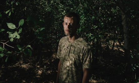Volodymyr Prysiazhniuk stands in a wooded area under the shade of leafy green trees. He wears a camouflage T-shirt and is looking at the camera