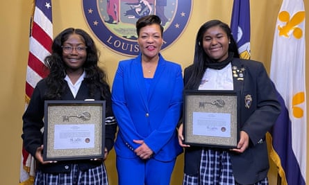 Ne’Kiya Jackson holding a picture-framed key standing in a row next to LaToya Cantrell and Calcea Johnson, who is also holding a picture-framed key, with various flags and a Louisiana state seal in the background. 