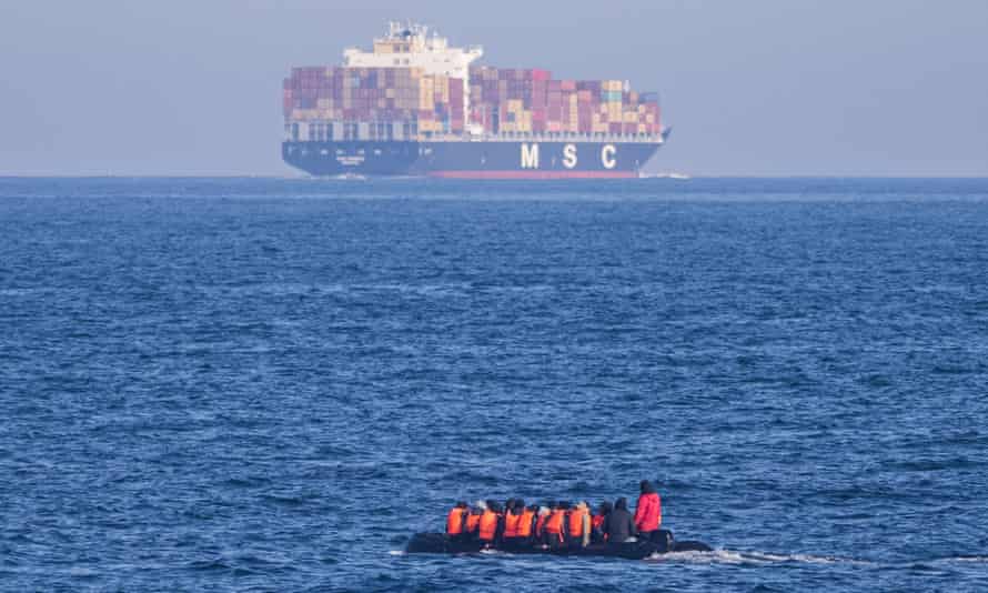 Personas con chalecos salvavidas naranjas sentadas en un bote pequeño y a poca distancia de un enorme buque de carga 