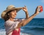 Senior woman taking a selfie on the beach with straw hat