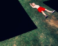 A young shirtless boy in red shorts lies on grass with arms stretched above his head