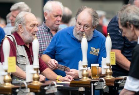 Men stand at a bar at the festival with drinks in front of them; they are all grey-haired and the two at the front have big white beards.