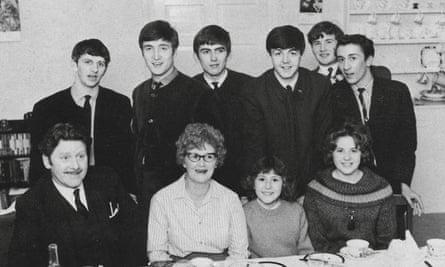 گروه بیتلز با چند مرد جوان دیگر پشت سر یک مرد، یک زن، یک زن جوانتر و یک کودک نشسته پشت میز ناهارخوری ایستاده اند.