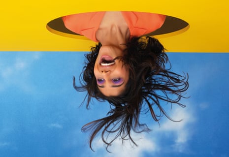 Woman in orange shirt with head through circular hole in yellow board, against blue sky background