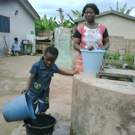 A young African girl, with very short hair, pours water from one bucket into another as a woman watches