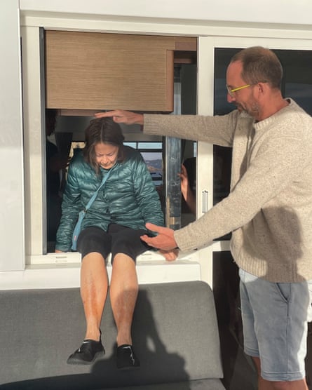 An older woman is helped out of a window on a catamaran by a man standing alongside her.