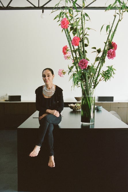 A woman wearing a giant necklace sits on a counter next to a towering floral arrangement