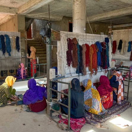 Women with covered heads sit in rows at three upright looms, while others crouch overstretched material in the middle of the workspace.