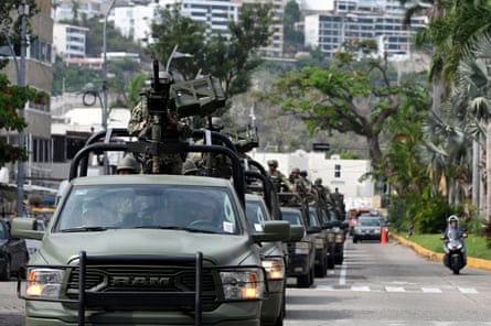 A queue of army trucks, each with a gunner on top, drive down a street
