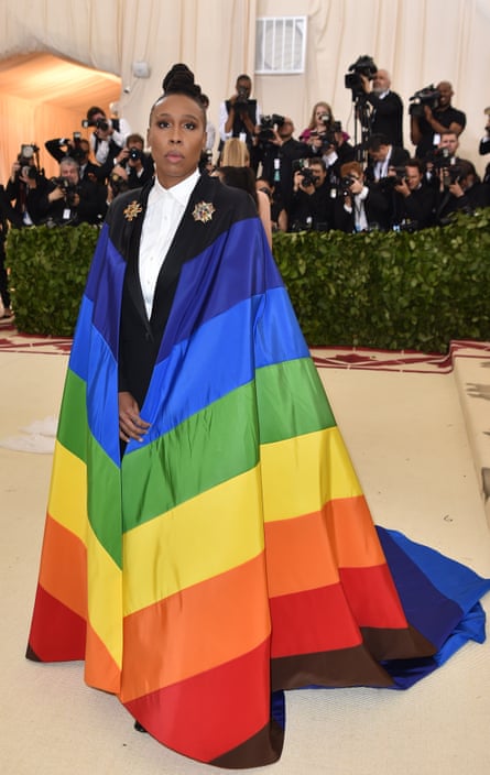 Lena Waithe standing in front of photographers wearing suit with long rainbow cape attached to shoulders.