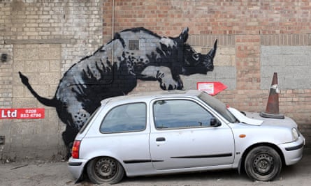 A large image of a rhino on a red-brick wall. The rhino appears to be clambering on top of a silver Nissan Micra parked next to the wall.
