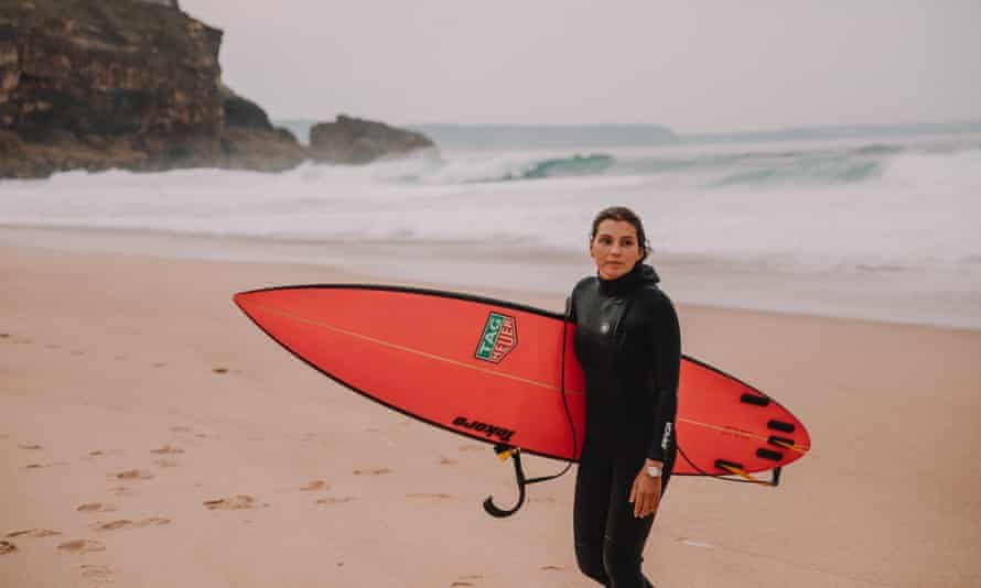 Maya Gabeira in her wetsuit, carrying her surfboard on a beach