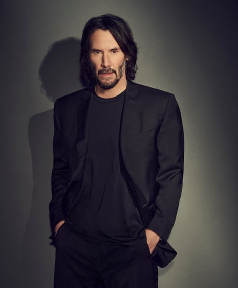 Keanu Reeves in a black suit and black t-shirt, looking at the camera, hands in pockets