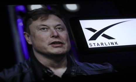 Elon Musk stands to one side of a screen displaying the Starlink logo