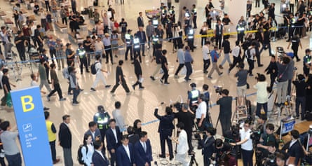 Korean passengers walking through an airport arrivals hall with rows of photographers and camera operators on either side