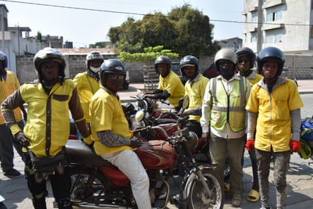 A group of moto-taxi drivers wearing helmets and yellow shirts gather around one driver sitting on a bike. 