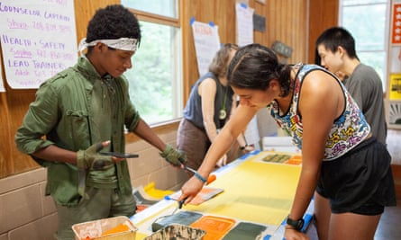 Inside a wood-panelled room, students in pairs stand across a table from each other painting signs. In the foreground a Black teenage boy wearing all olive green with a white bandana holding back his black hair paints with a white-presenting girl wearing a brightly colored Keith Haring tank top and black shorts, her medium-length brown hair pulled back in two short pigtails.