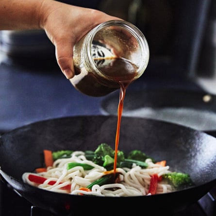 A jar of brown-coloured sauce being poured into a wok filled with udon and Asian greens.