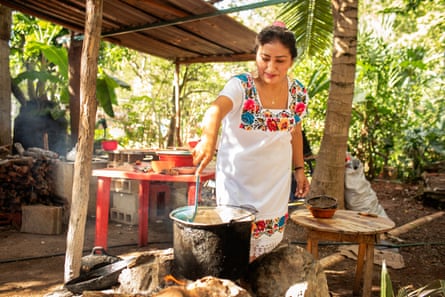 A woman in an embroidered dress stirs a large steaming cooking pot in an outdoor kitchen surrounded by greenery