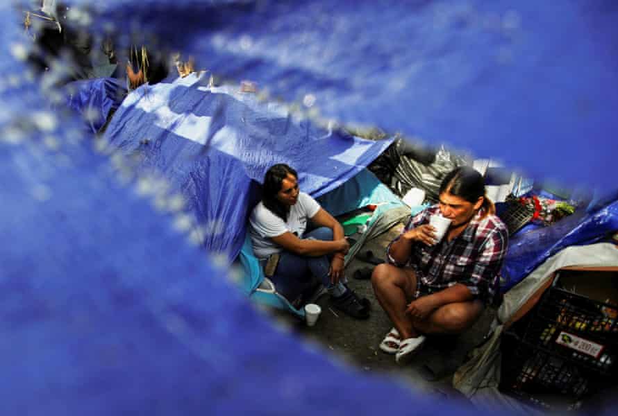 Two women drink coffee while sitting in the shade of torn blue tarps.