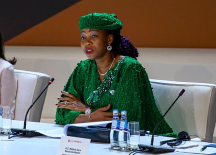 A middle-aged African woman in an elaborately beaded green dress and hat sits at a table talking