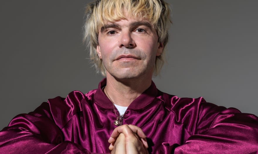 Tim Burgess in a shiny dark pink jacket, hands linked together in front of his chest